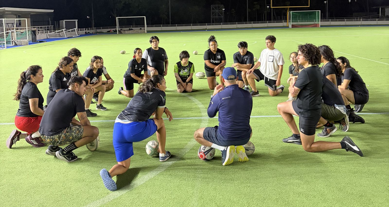 El equipo, sentado en círculo sobre la cancha, escucha a la coach en un entrenamiento nocturno